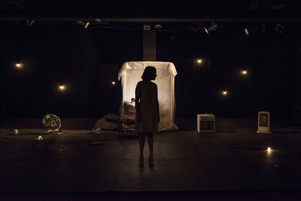 Escena en penumbras, con algunas lámparas que apenas se ven encendidas en sobre el fondo. En el centro un colchón parado, apoyado sobre la pared del fondo, iluminado más que el resto de la escena, en el frente, se ve la silueta en sombras de una mujer de pie mirando hacia un lado.