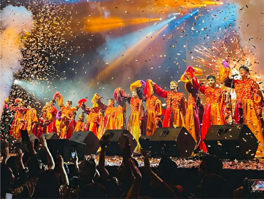 Murga La Zafada, foto frontal de un momento de la despedida en el Teatro de Verano Ramón Collazo, trajes de raso naranjas y rojos con gorros estilo bufon 23 niños saludan con la mano alzada y el gorro en la mano