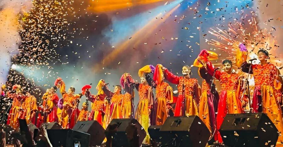 Murga La Zafada, foto frontal de un momento de la despedida en el Teatro de Verano Ramón Collazo, trajes de raso naranjas y rojos con gorros estilo bufon 23 niños saludan con la mano alzada y el gorro en la mano